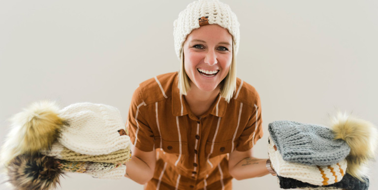 Woman in a knit hat standing with a pile of knit hats on each hand.