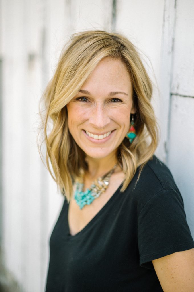 Marji Guyler-Alaniz smiling with her back against a white painted wood wall.
